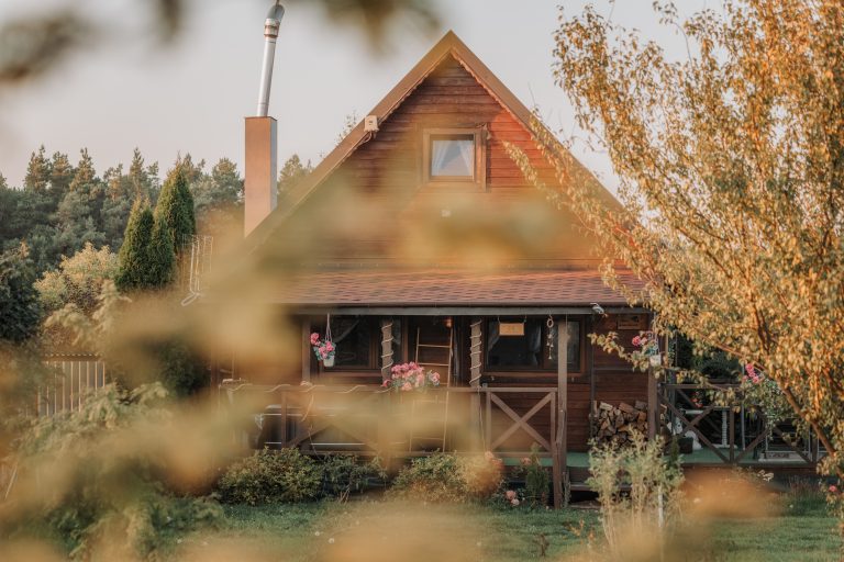 Drewniany domek całoroczny Wichrowa Chata w Jońcu otoczony roślinnością i kwiatami, na tle lasu i zielonego ogrodu, widziany przez gałęzie drzew.